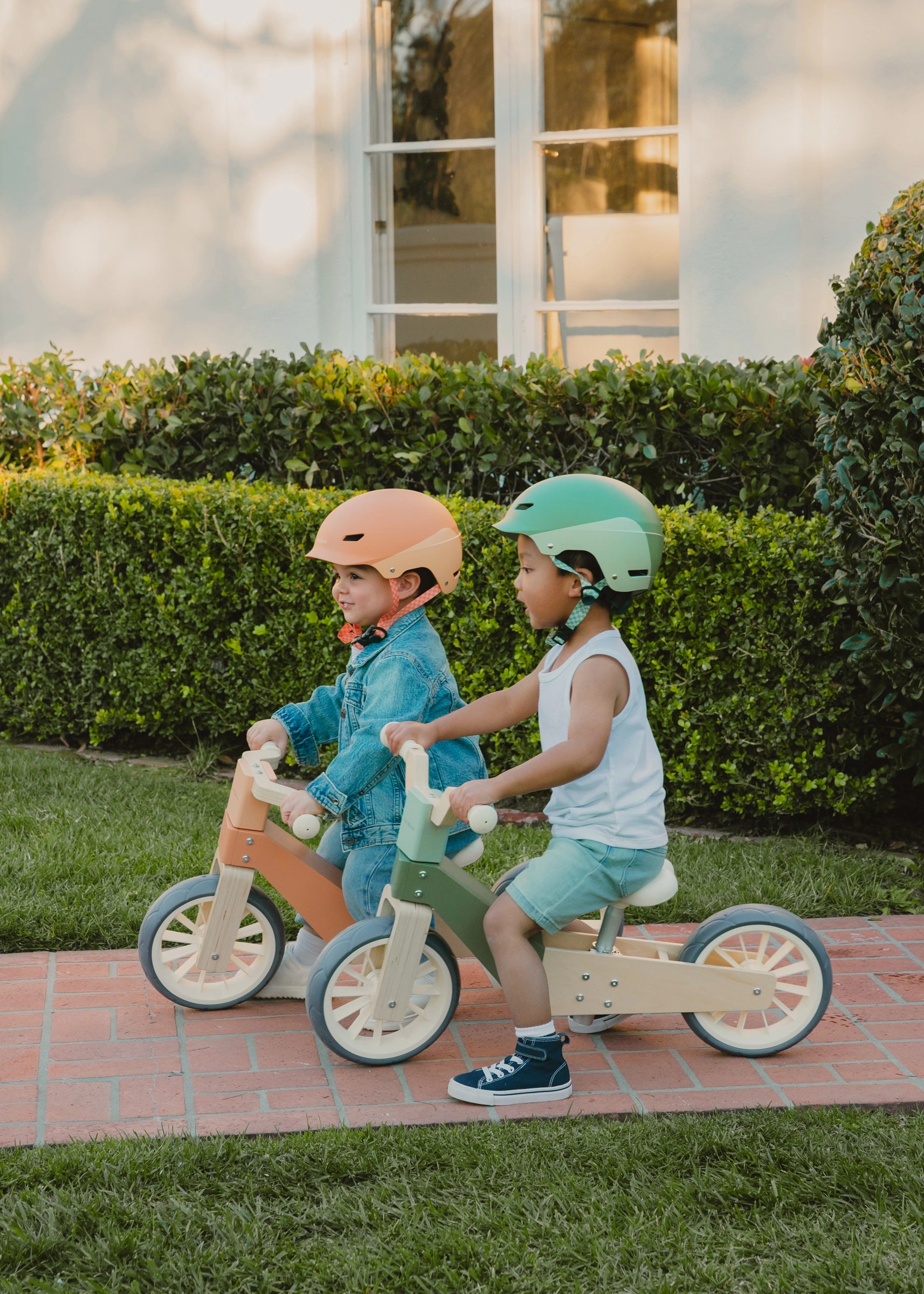 Two children riding balance bikes on a sidewalk with a house and greenery in the background.