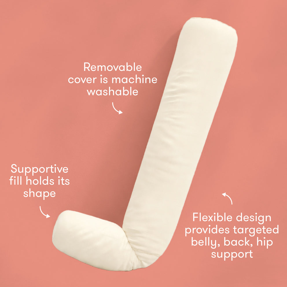 An off-white curved maternity support pillow on a solid coral pink background, surrounded by three white text callouts explaining its features: 'Removable cover is machine washable', 'Supportive fill holds its shape', and 'Flexible design provides targeted belly, back, hip support'.