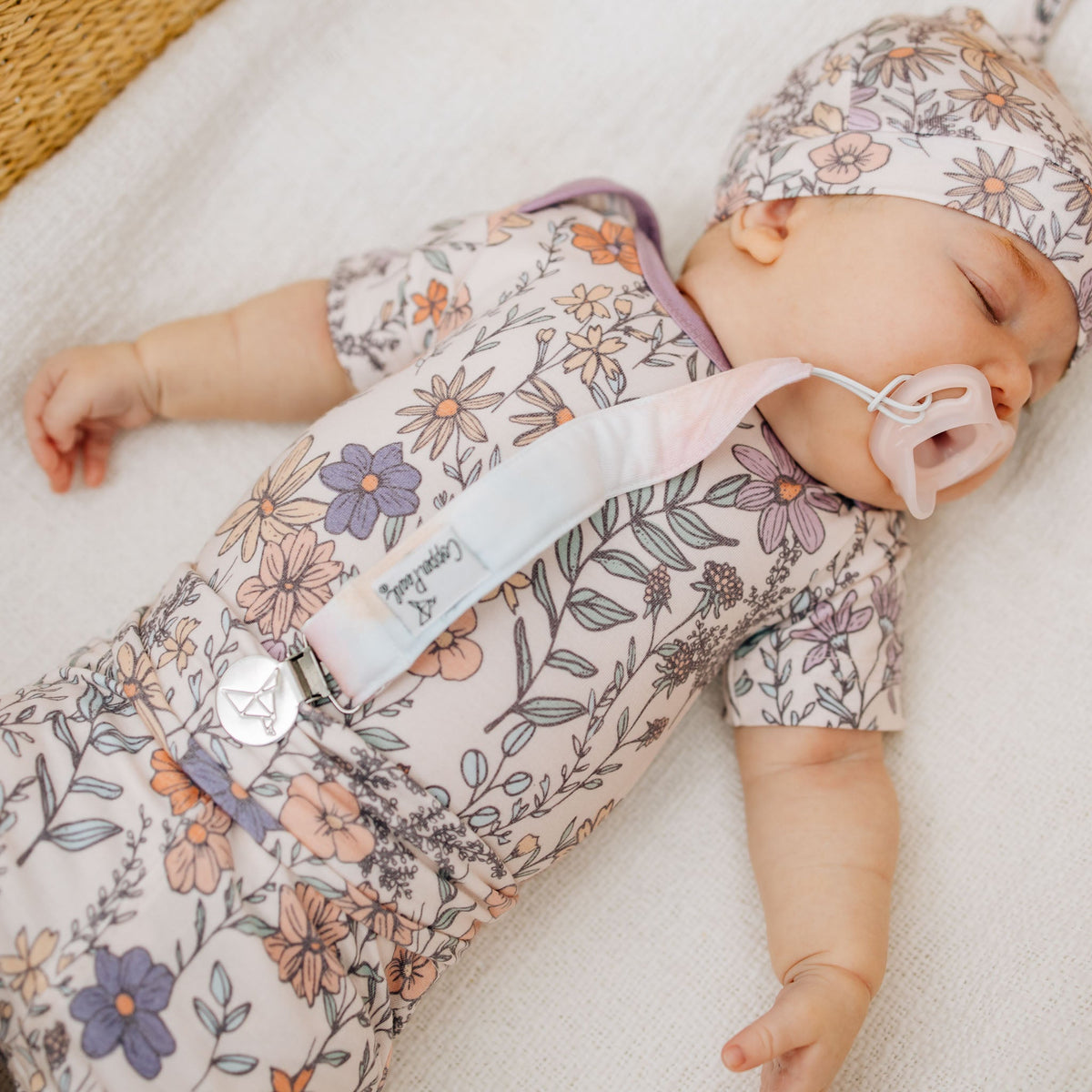 Baby sleeping in a floral outfit with a pacifier, on a soft surface.