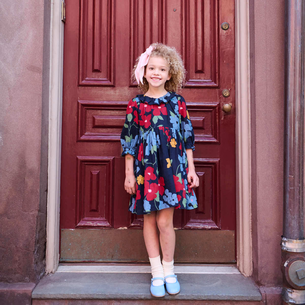 A young girl with curly blonde hair stands on a doorstep in front of a dark red wooden door, wearing a navy blue floral dress with red, yellow, and light blue flowers, white socks, and light blue shoes.
