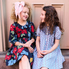 Two young girls sitting side-by-side wearing vintage-style floral dresses. The girl on the left has curly blonde hair and wears a navy blue dress with a red, blue, and yellow floral pattern, along with a pink bow headband.