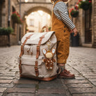 Child in mustard yellow overalls standing next to a large beige backpack with a plush toy on a cobblestone street.