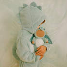 Child wearing a green dinosaur costume holding a matching plush toy against a soft, neutral background.