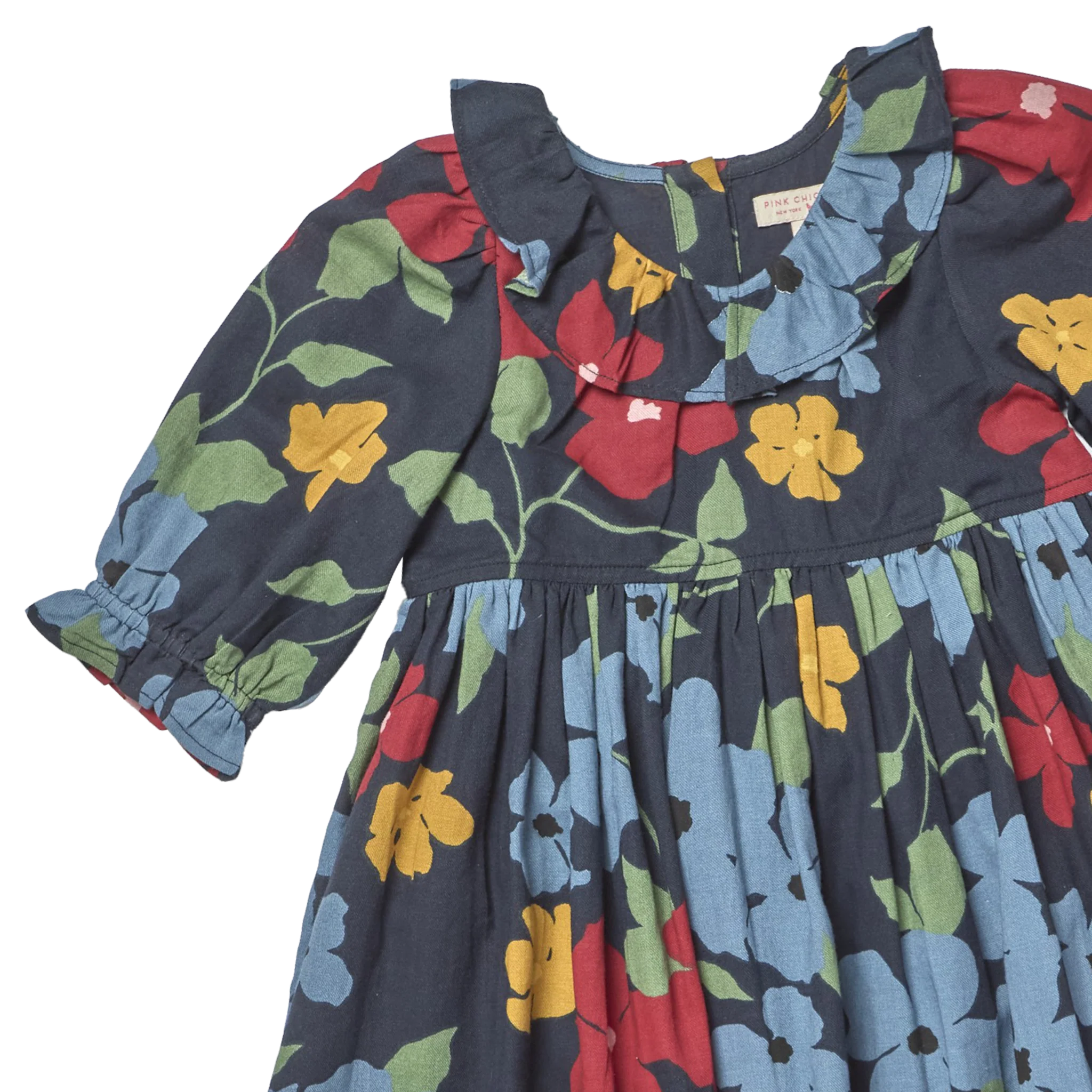 A close-up of a children's dress with a dark navy blue base and a colorful floral print featuring red, yellow, and light blue flowers with green leaves. The dress has a ruffled collar, long sleeves with elastic cuffs, and a gathered waist.