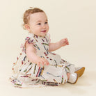 A baby girl sitting and smiling, wearing a white ruffled dress with a colorful crayon print and silver shoes, set against a solid cream background.