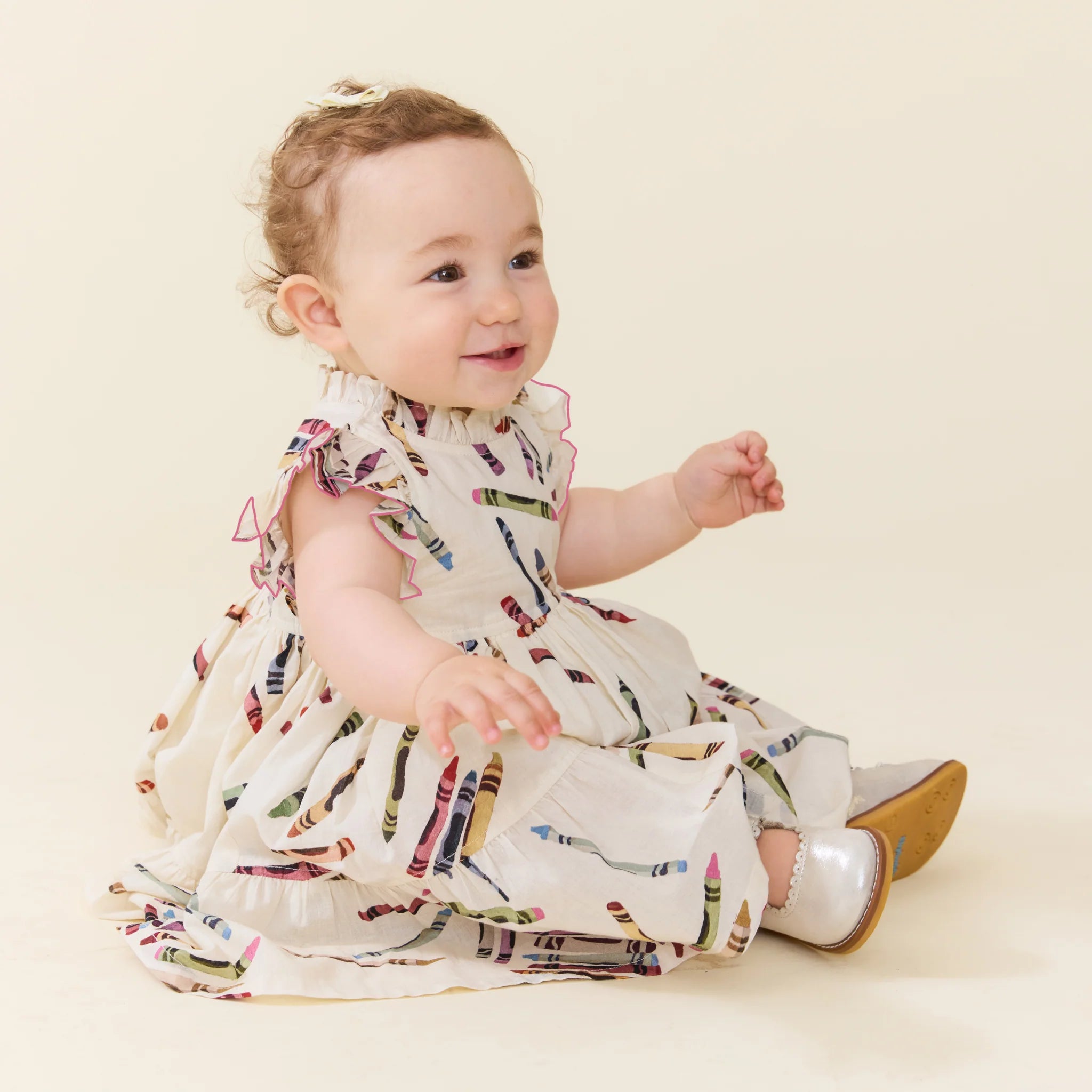 A baby girl sitting and smiling, wearing a white ruffled dress with a colorful crayon print and silver shoes, set against a solid cream background.