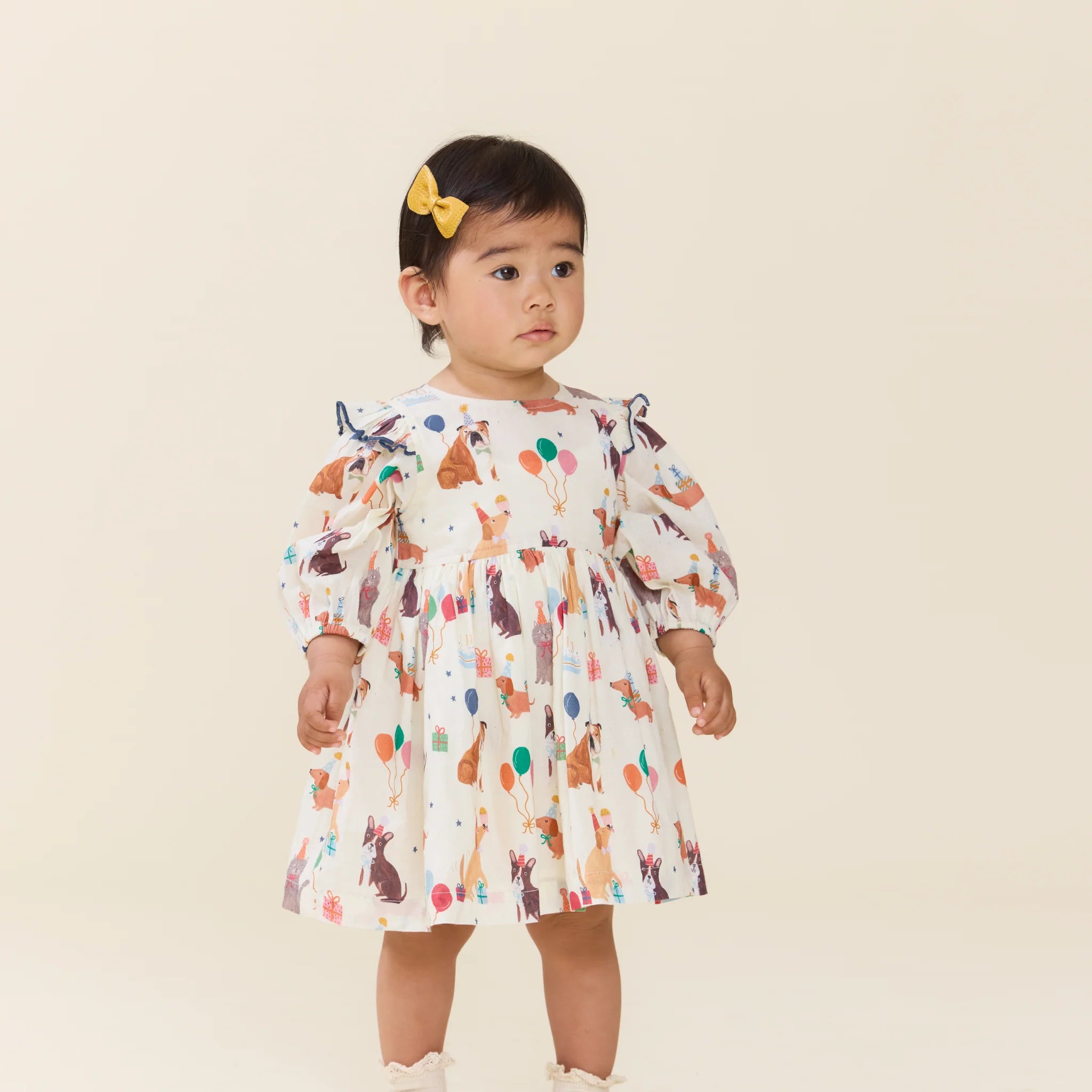 A toddler girl wearing a white long-sleeve dress with a colorful dog and balloon print, standing against a solid light beige background. She has a yellow bow in her hair.