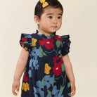 A toddler girl wearing a navy blue floral dress with ruffled sleeves, photographed against a plain beige background. She has a yellow bow in her hair.