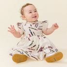 A baby sitting and smiling, wearing a white dress with a colorful crayon print and tan sole shoes, photographed against a plain cream background.