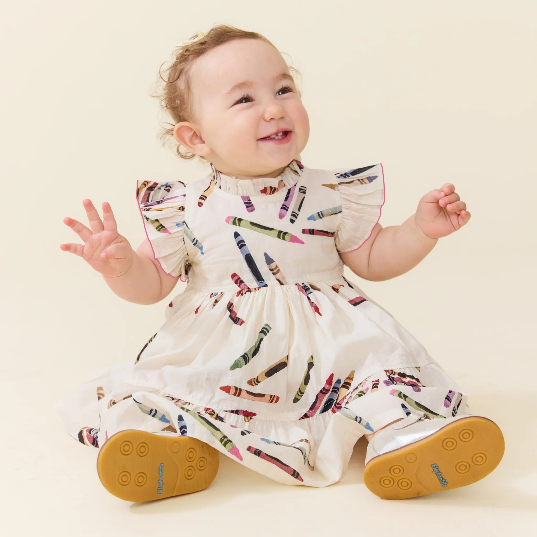 A baby sitting and smiling, wearing a white dress with a colorful crayon print and tan sole shoes, photographed against a plain cream background.