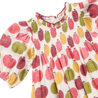A close-up of a children's dress with a white base and an all-over apple print in pink, red, yellow, and green. The dress features a smocked bodice, ruffled neckline, and short puff sleeves.