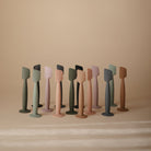 A set of ten silicone kitchen spatulas in muted earthy colors, including sage green, dusty pink, terracotta, and charcoal, arranged in a row against a warm beige background.
