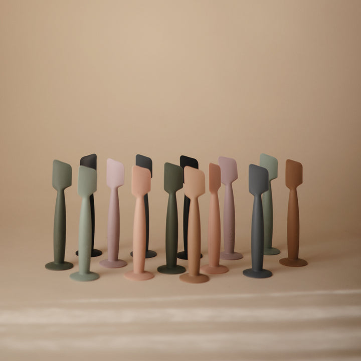 A set of ten silicone kitchen spatulas in muted earthy colors, including sage green, dusty pink, terracotta, and charcoal, arranged in a row against a warm beige background.