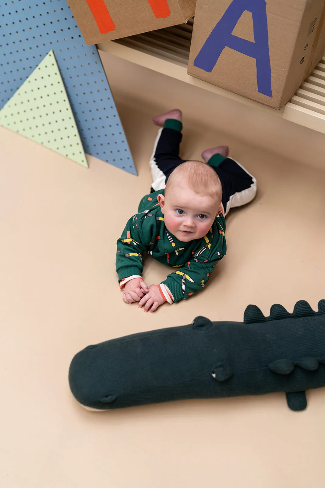 A baby wearing a green patterned long-sleeve baby jumpsuit lies on their stomach on a beige floor, with a dark green plush crocodile toy in the foreground and colorful geometric wall decorations in the background.
