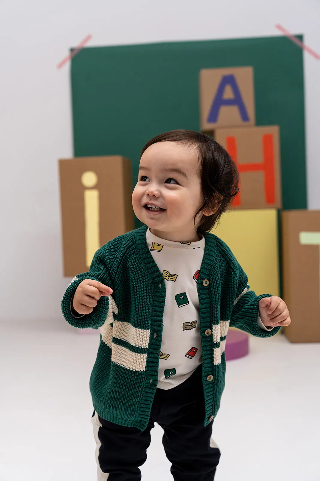 A smiling toddler wearing a green knit cardigan with cream stripes over a white patterned top and dark pants, standing in front of a green backdrop with colorful letter blocks.