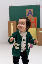 A smiling toddler wearing a green knit cardigan with cream stripes over a white patterned top and dark pants, standing in front of a green backdrop with colorful letter blocks.