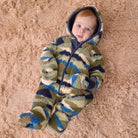 A baby lying on a beige shag rug, wearing a plush hooded onesie with a camouflage pattern in olive green, navy blue, and cream.