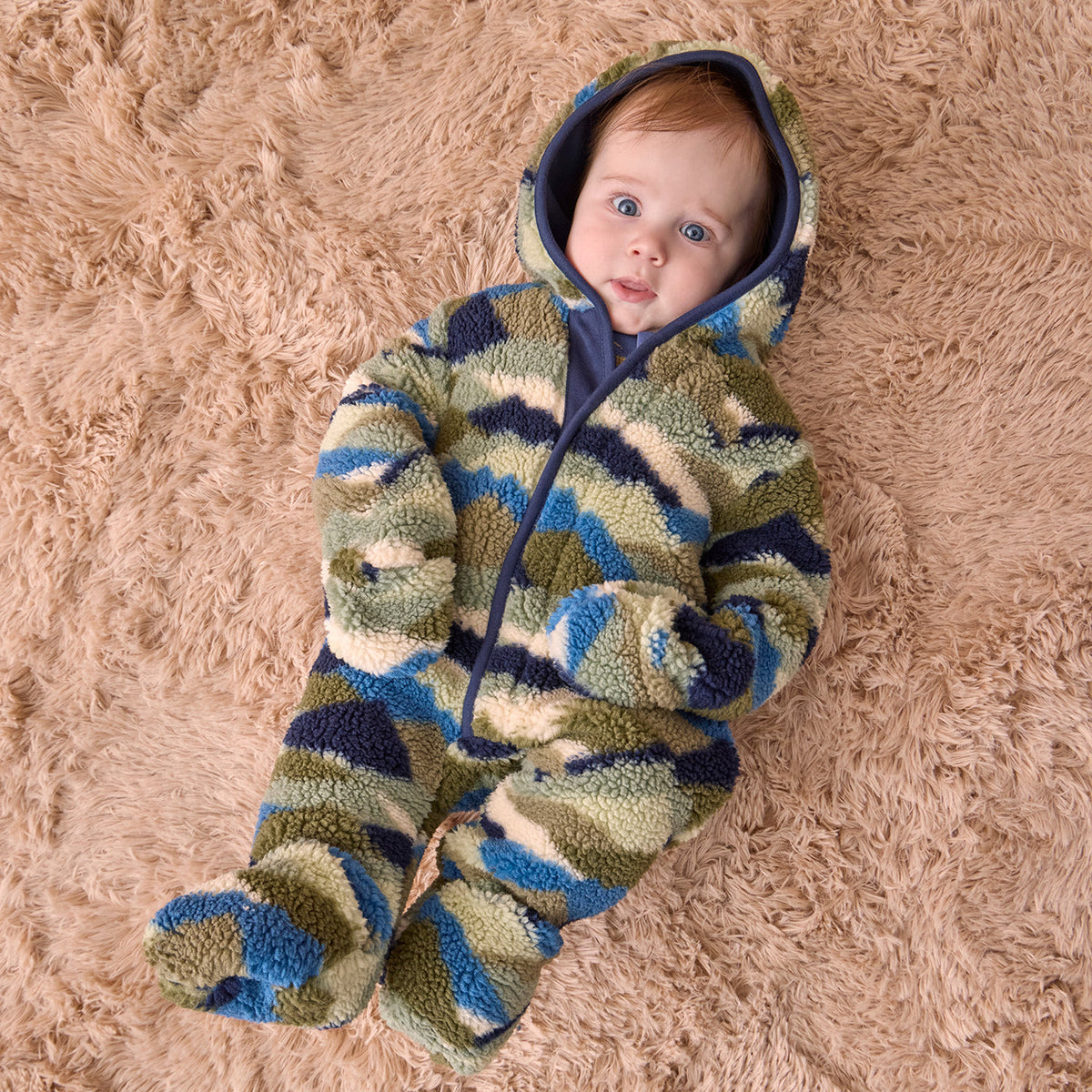 A baby lying on a beige shag rug, wearing a plush hooded onesie with a camouflage pattern in olive green, navy blue, and cream.