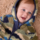 A baby lying on a beige shag rug, wearing a plush hooded onesie with a camouflage pattern in olive green, navy blue, and cream.