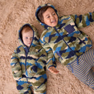 Two children lying on a fluffy beige rug wearing matching hooded one-piece outfits with a blue, green, and beige camouflage pattern. The larger child is smiling and wearing striped pants, while the smaller child looks toward the camera.