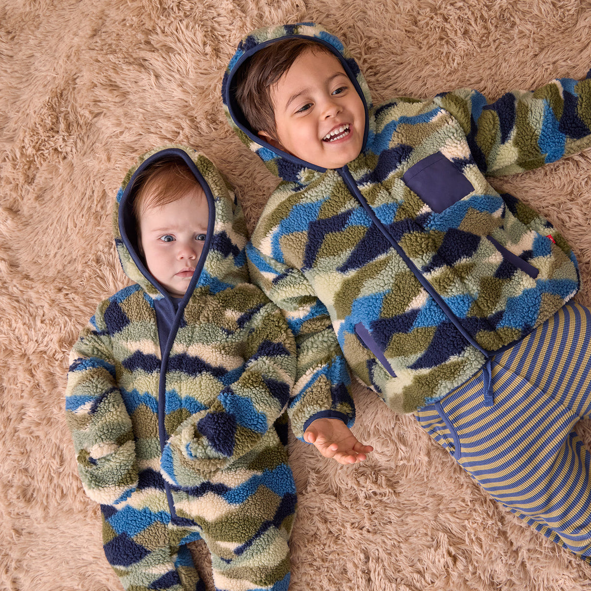 Two children lying on a fluffy beige rug wearing matching hooded one-piece outfits with a blue, green, and beige camouflage pattern. The larger child is smiling and wearing striped pants, while the smaller child looks toward the camera.