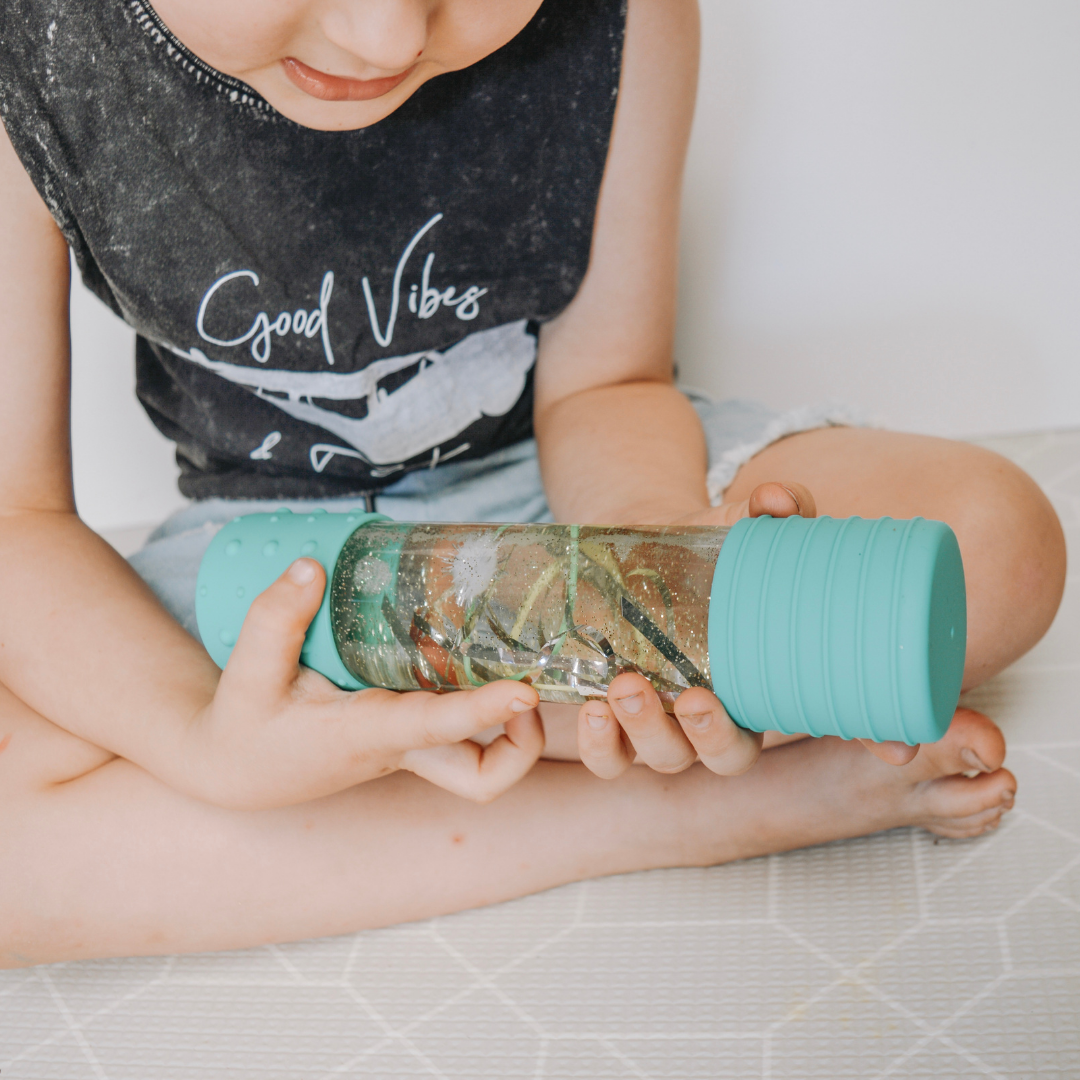 Child holding a turquoise water bottle with a clear lid on a light-colored floor.