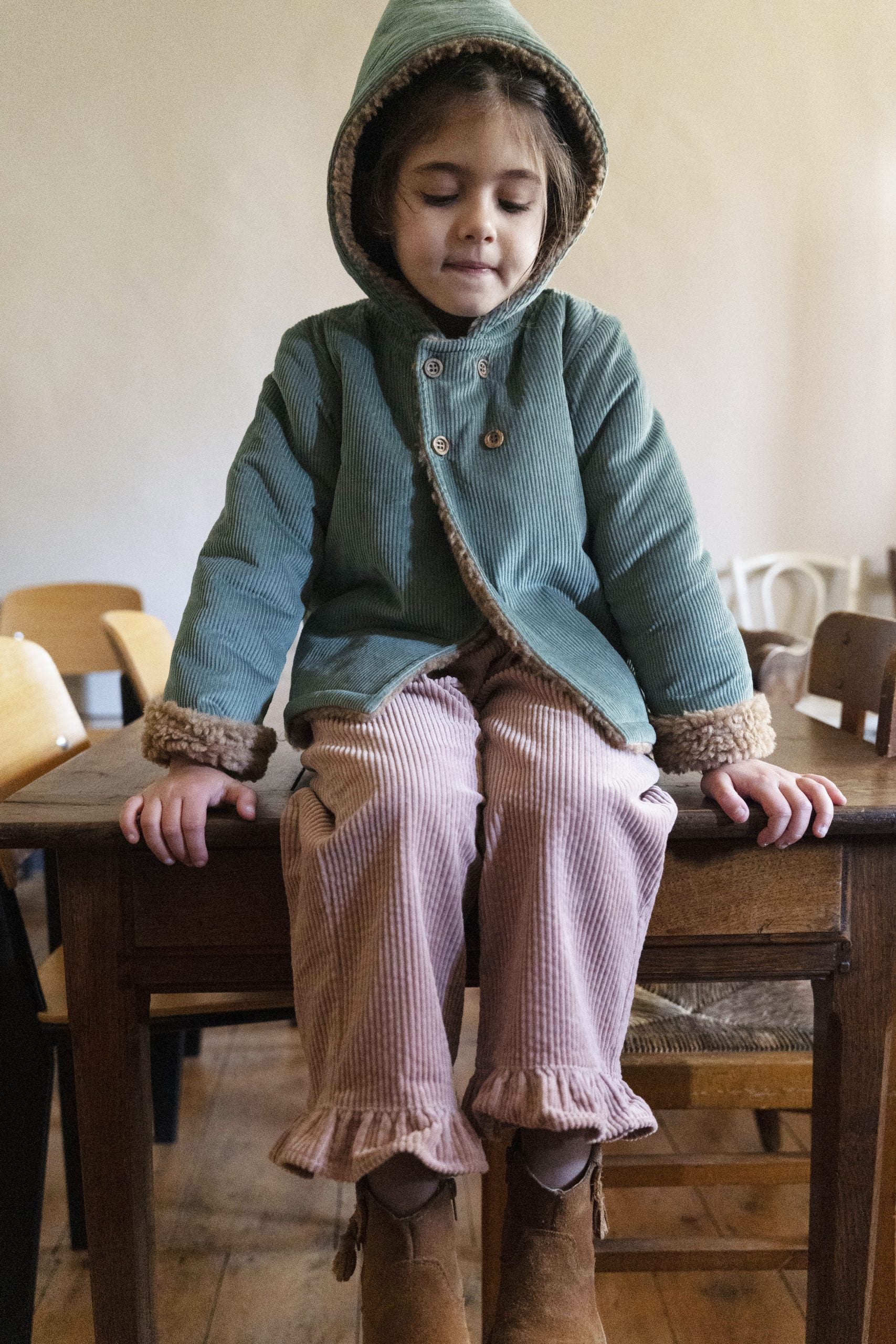 A young child wearing a muted sage green hooded jacket with brown faux-fur trim and pink ribbed trousers, sitting on a rustic wooden table in a warm indoor setting.