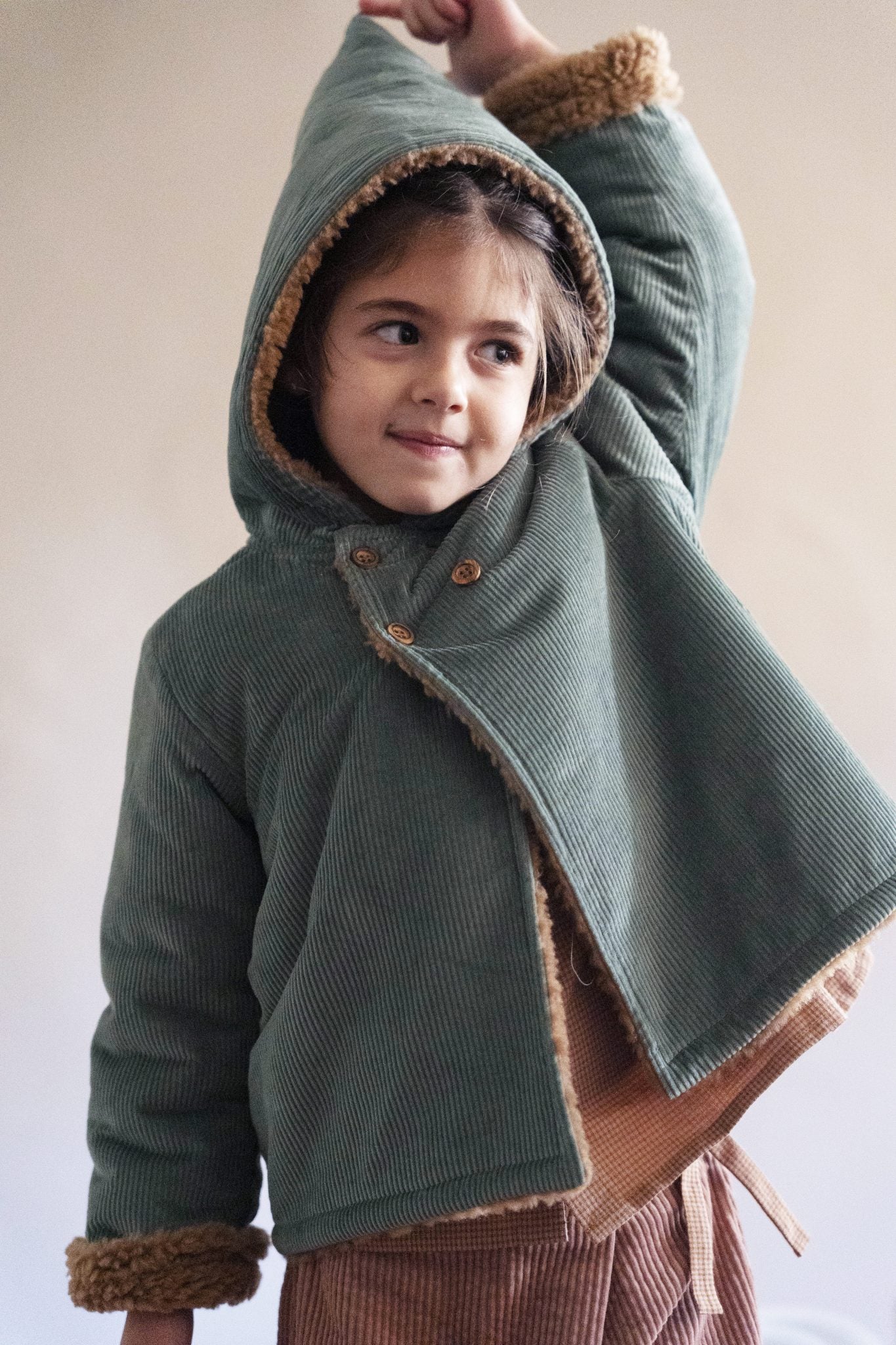 A young child wearing a sage green corduroy hooded jacket with brown faux-fur trim, posed against a soft beige background.