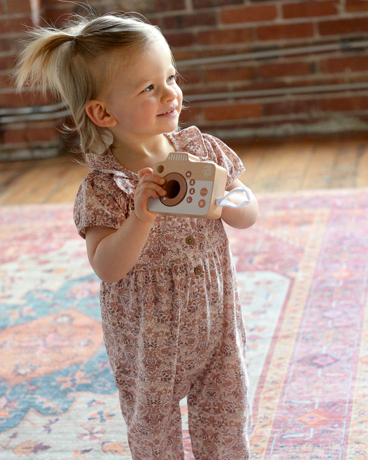 Child holding a toy camera on a patterned rug with a brick wall background