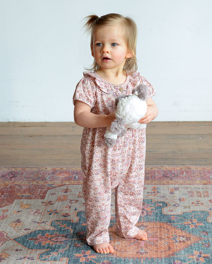 Young child in a floral outfit holding a plush toy on a patterned rug.