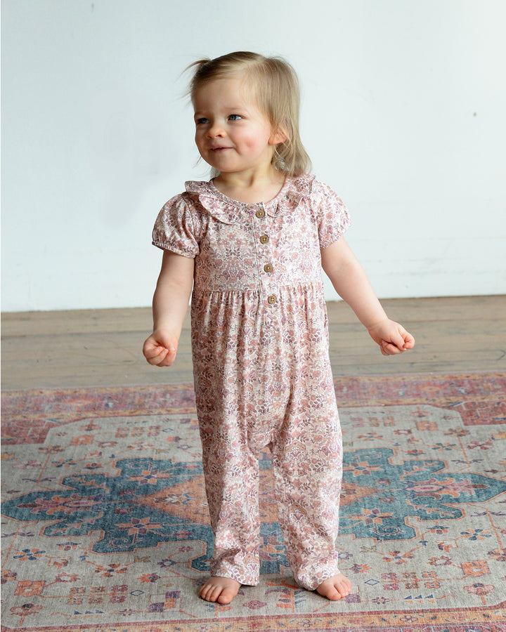 Young child wearing a floral romper standing on a patterned rug.