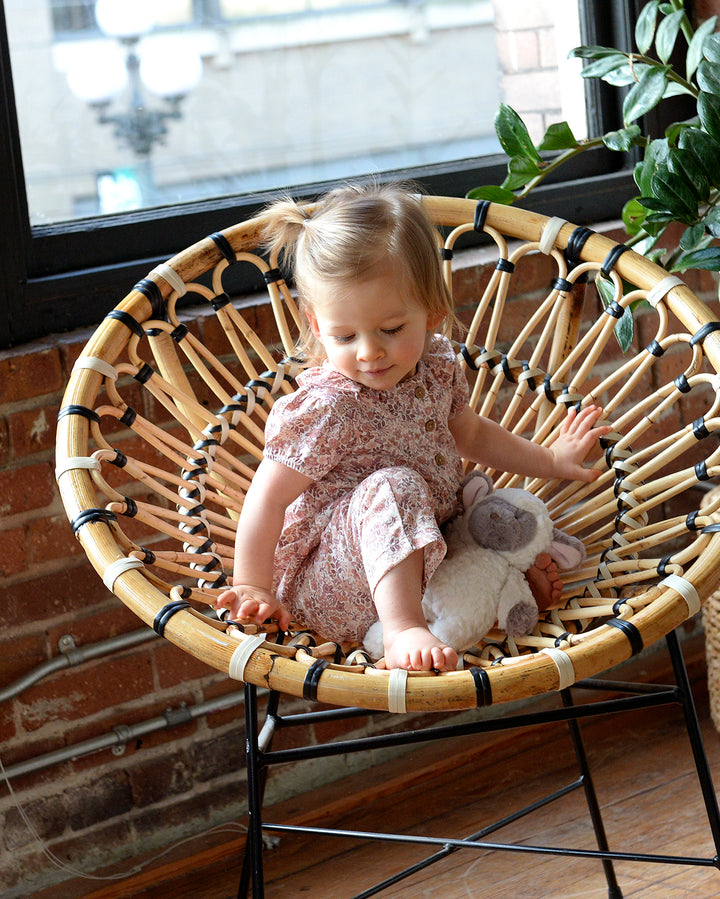 Child sitting in a wicker chair holding a stuffed animal, with a plant and window in the background.