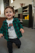 Child wearing a green plaid shirt over a white t-shirt with a graphic design, standing in a room.