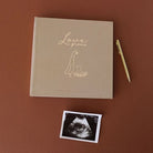 A square tan keepsake book with gold foil lettering reading 'Love grows' and a simple baby silhouette, displayed on a warm brown background with a gold pen and an ultrasound photo print below it.