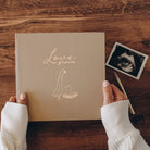 Book titled 'Love Grows' with a hand holding it on a wooden surface, next to an ultrasound photo.