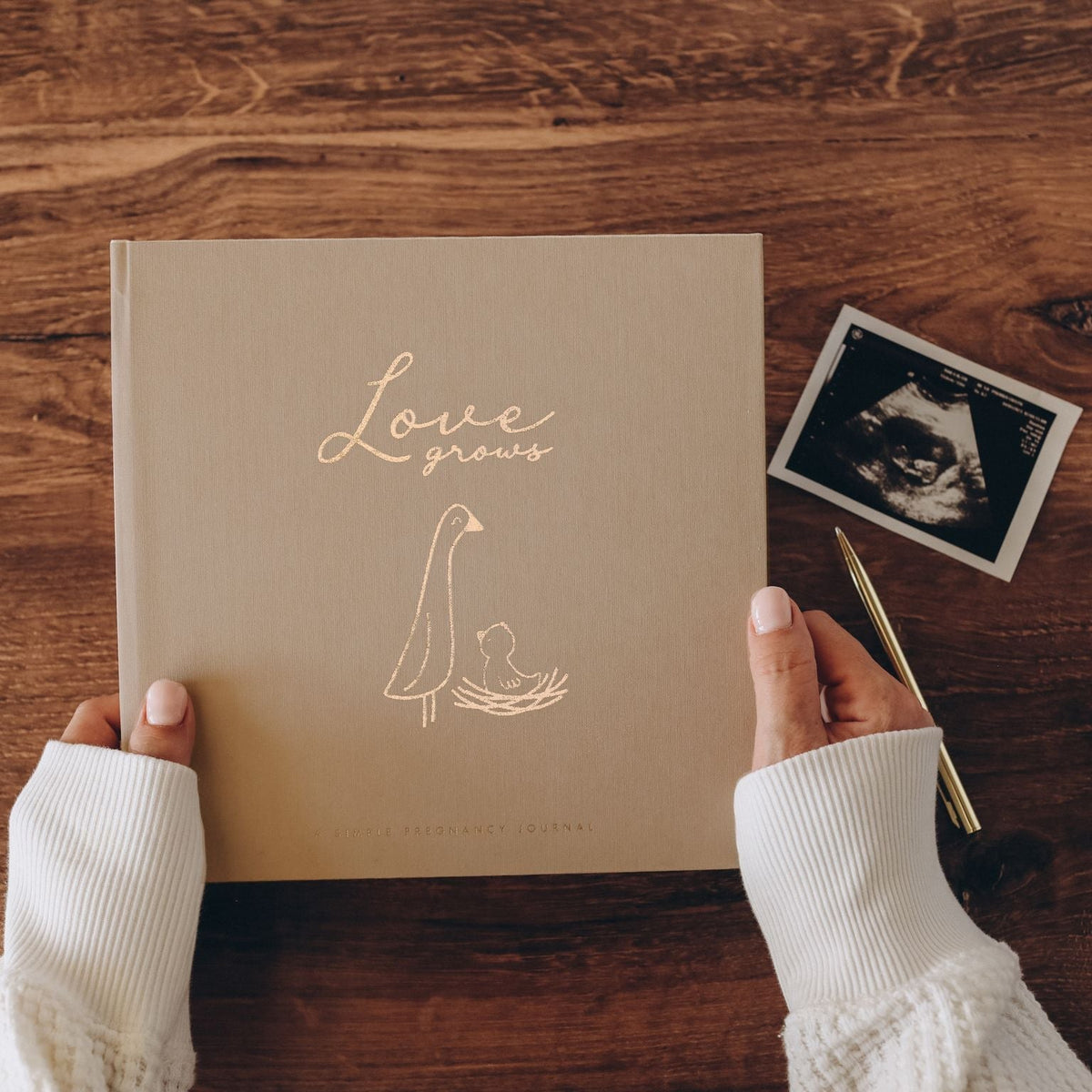 Book titled 'Love Grows' with a hand holding it on a wooden surface, next to an ultrasound photo.