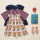 A styled flat-lay of a children's outfit set on a warm cream background, including a light pink dress with a colorful milk carton print, a pink-and-purple gingham long-sleeve shirt, and a navy blue puffed-sleeve top. Props include a red apple, a stack of blue books, a small green box, and pink shoes.