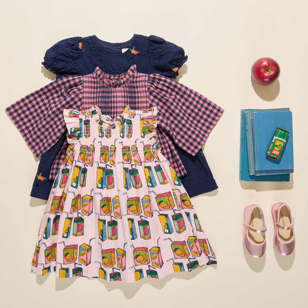 A styled flat-lay of a children's outfit set on a warm cream background, including a light pink dress with a colorful milk carton print, a pink-and-purple gingham long-sleeve shirt, and a navy blue puffed-sleeve top. Props include a red apple, a stack of blue books, a small green box, and pink shoes.