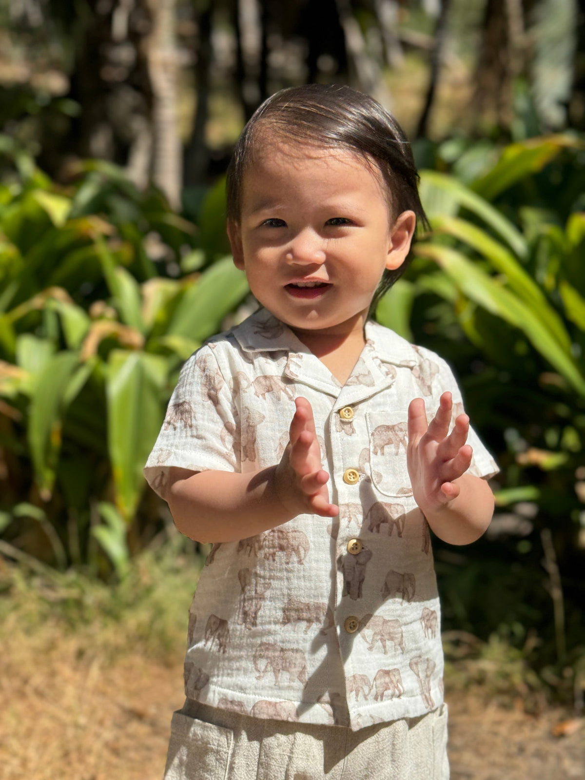 A toddler wearing a matching short-sleeve button-up shirt and shorts set in a light beige linen-like fabric with a brown elephant print, standing outdoors in front of lush green foliage.