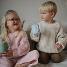 Two children sitting on a couch, one holding a green cup and the other a blue cup.