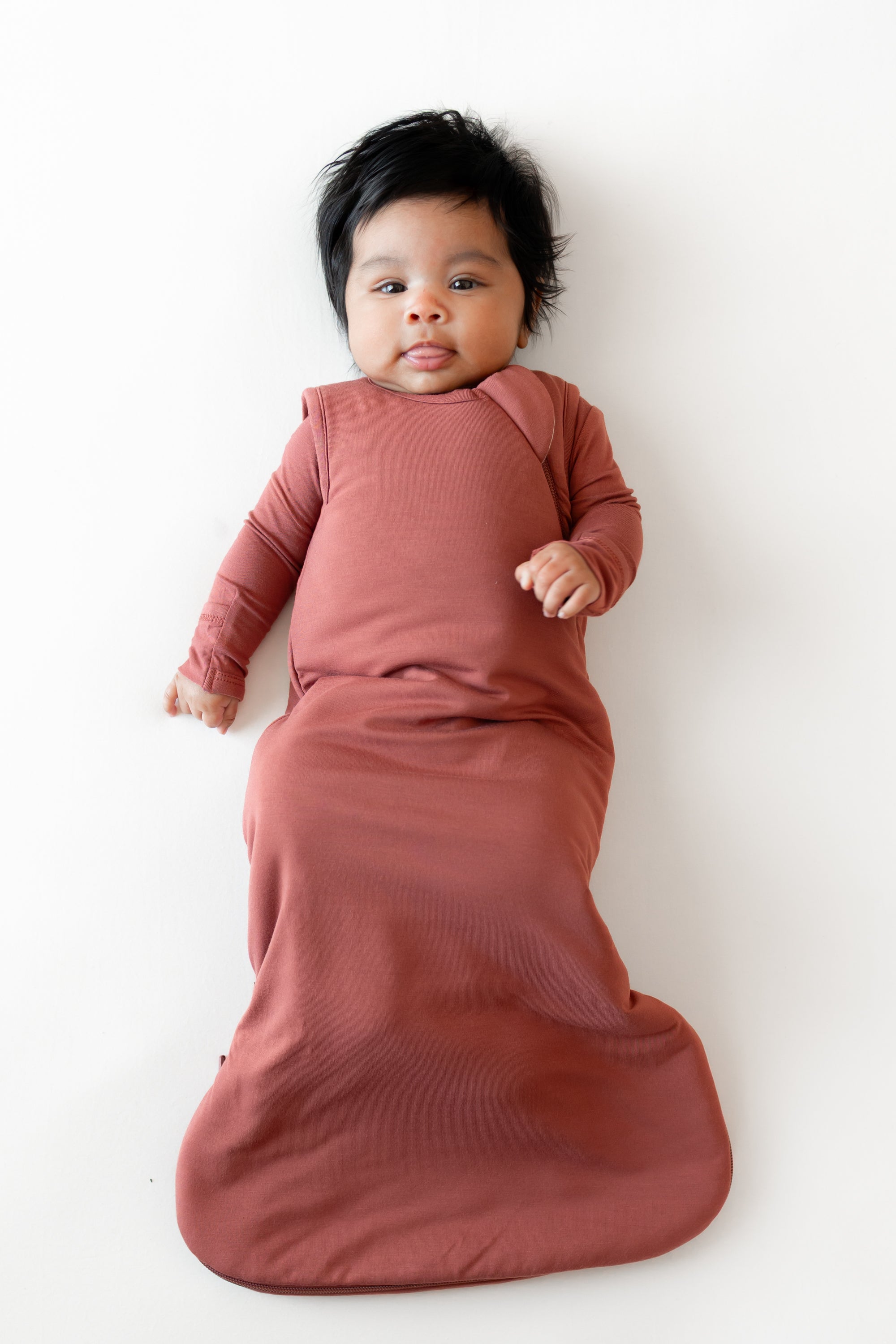 A baby lying on their back wearing a dusty rose long-sleeve swaddle blanket against a clean white background.
