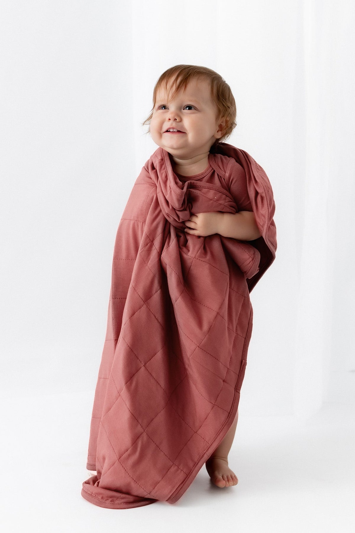 A toddler stands barefoot on a white studio background wearing a dusty rose short-sleeve bodysuit and holding a matching draped fabric sheet.