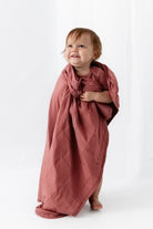 A toddler stands barefoot on a white studio background wearing a dusty rose short-sleeve bodysuit and holding a matching draped fabric sheet.