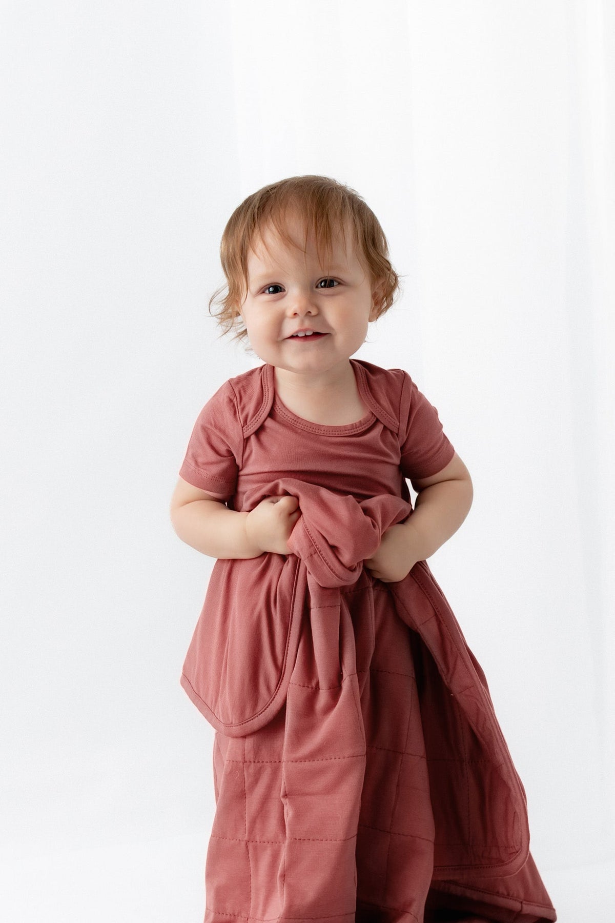A toddler with light brown hair stands against a white studio background wearing a dusty rose pink short-sleeve bodysuit and holding a matching quilted blanket.