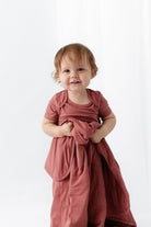 A toddler with light brown hair stands against a white studio background wearing a dusty rose pink short-sleeve bodysuit and holding a matching quilted blanket.