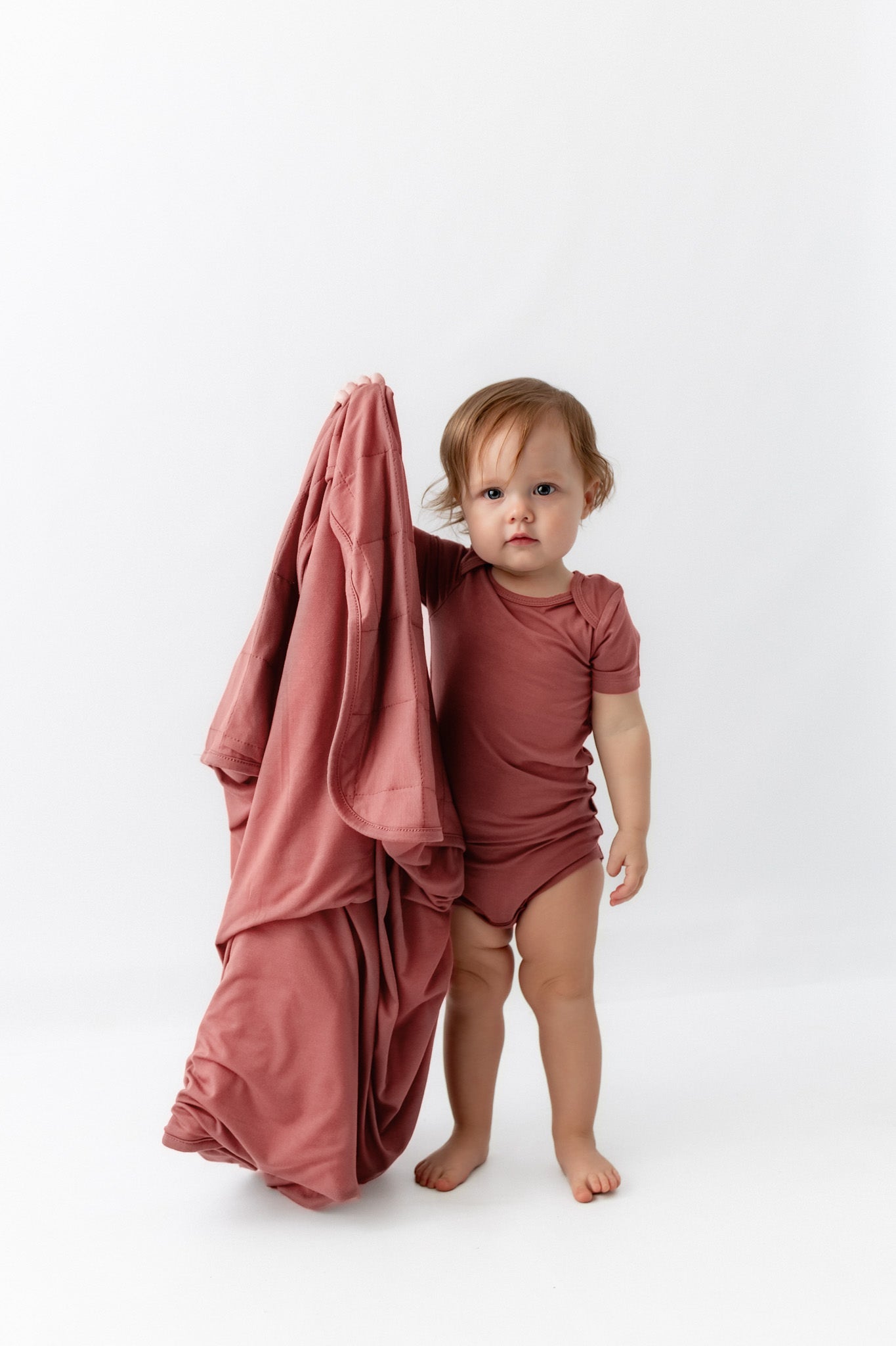 A toddler stands barefoot on a white studio background wearing a dusty rose short-sleeve bodysuit and holding a matching draped fabric sheet.