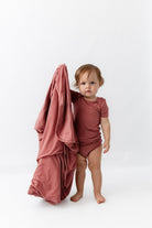 A toddler stands barefoot on a white studio background wearing a dusty rose short-sleeve bodysuit and holding a matching draped fabric sheet.