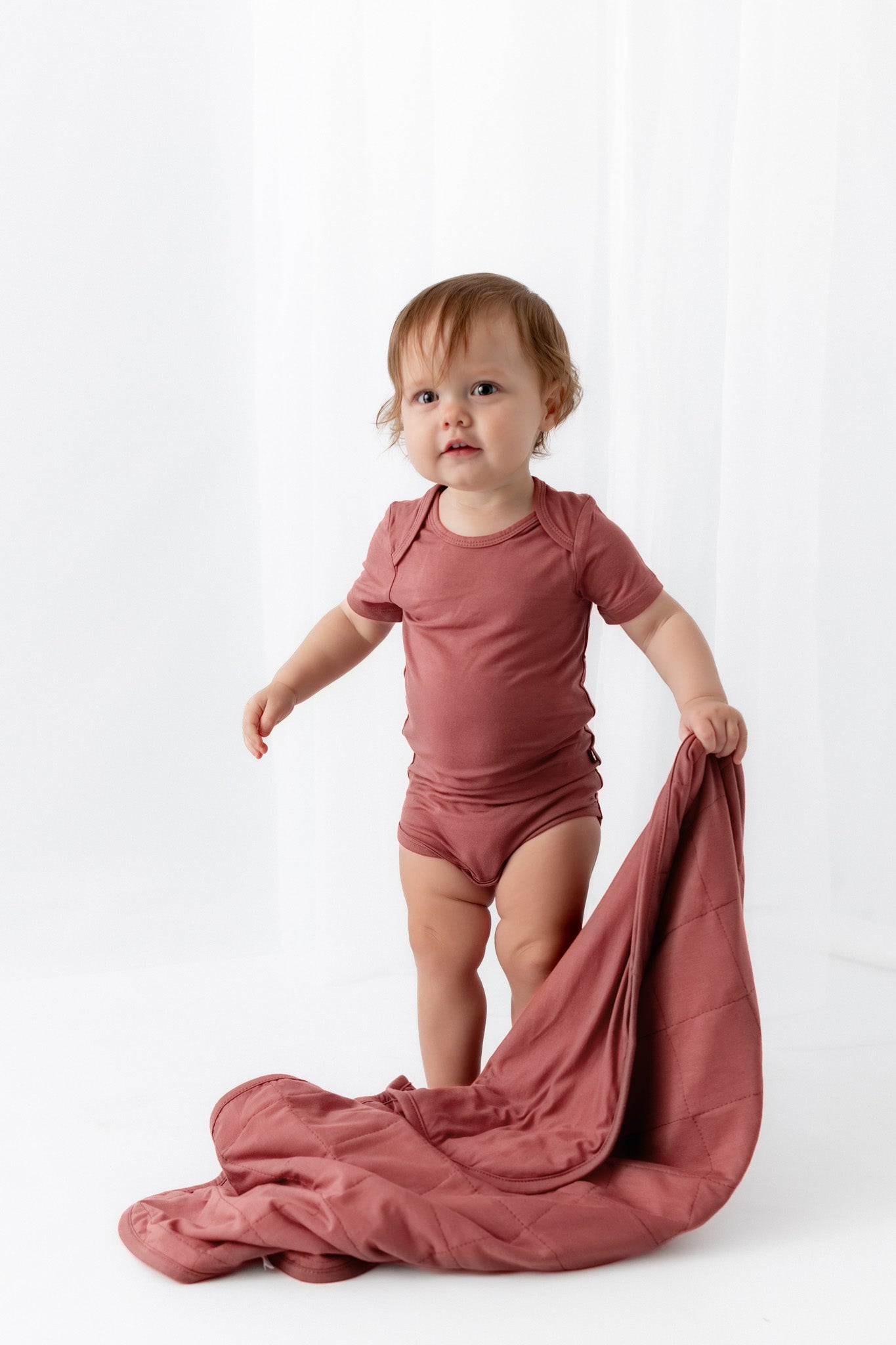 A toddler stands in a studio wearing a dusty rose short-sleeve bodysuit and holding a matching dusty rose sheet or blanket spread on the floor.