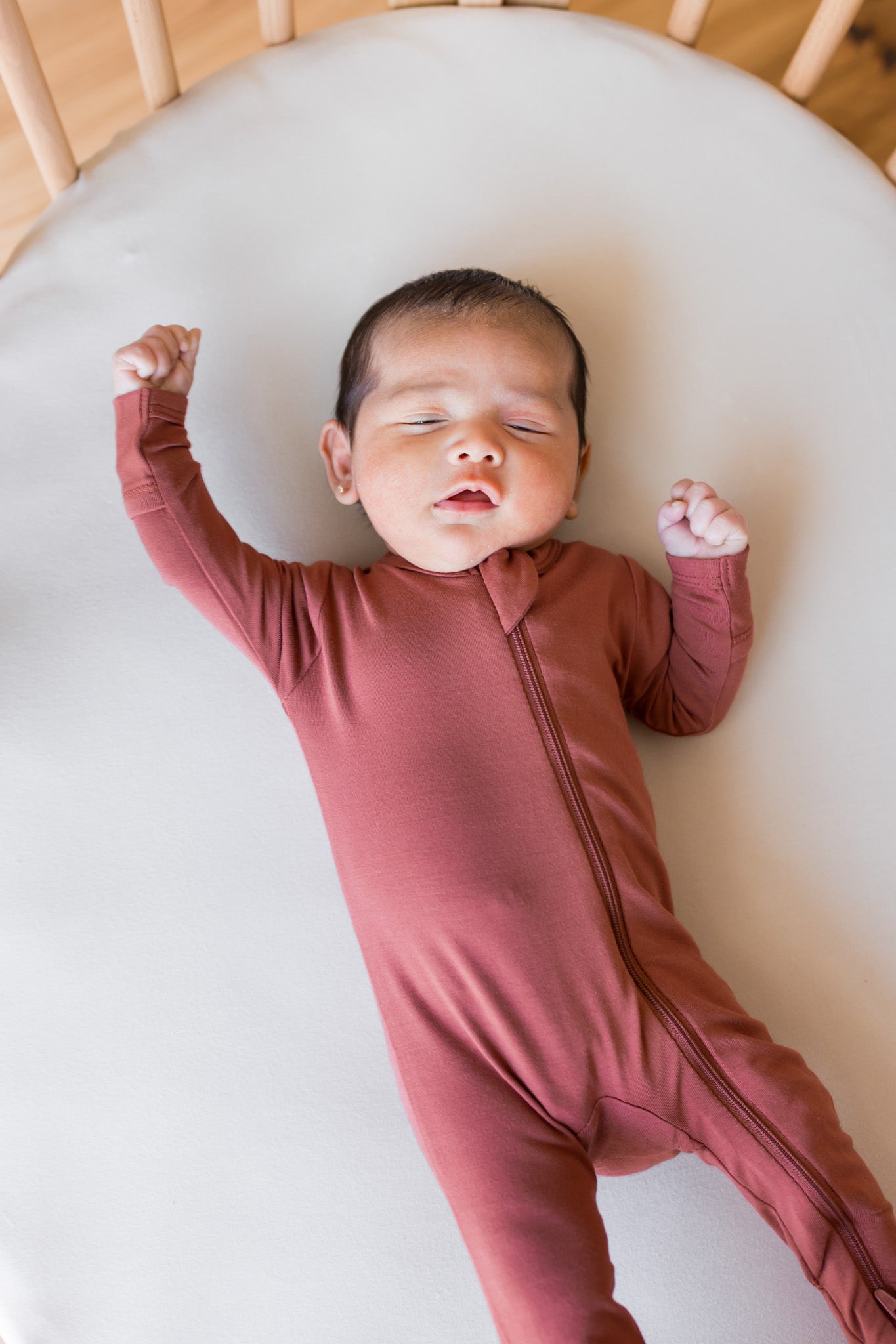 A newborn baby wearing a dusty rose long-sleeve zip-up footless onesie,  lying on a white oval mattress inside a wooden crib.