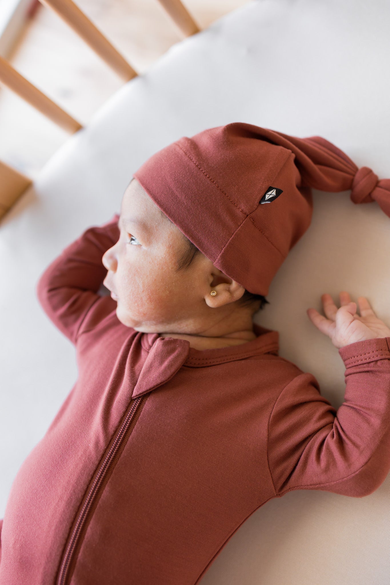 A newborn baby wearing a dusty rose long-sleeve zip-up footless onesie and knotted cap  lying on a white oval mattress inside a wooden crib.